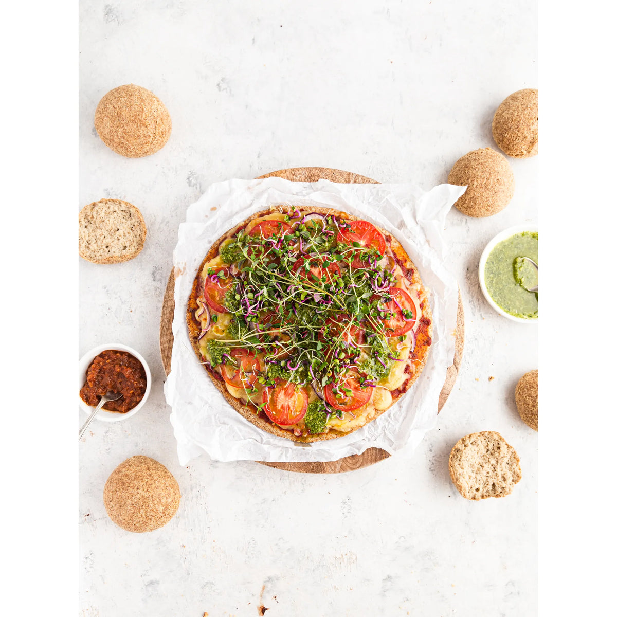 Vegetable  pizza with herbs on a white surface with crackers and a bread rolls and a small dish of pesto.