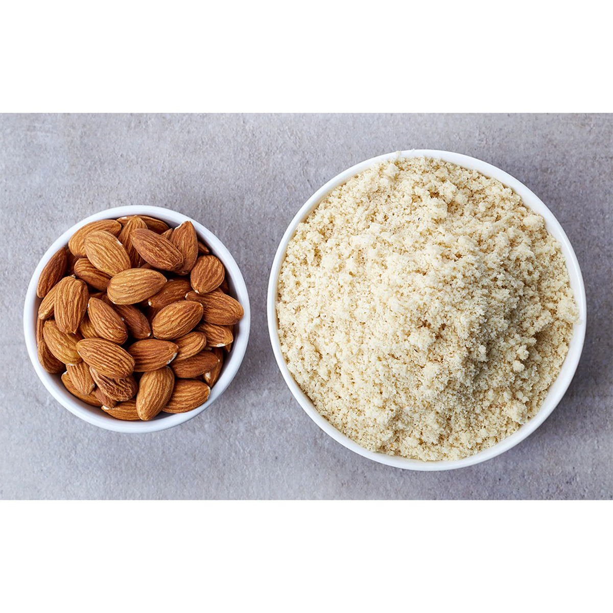 Two bowls, one with almonds and the other with almond flour, on a gray surface.