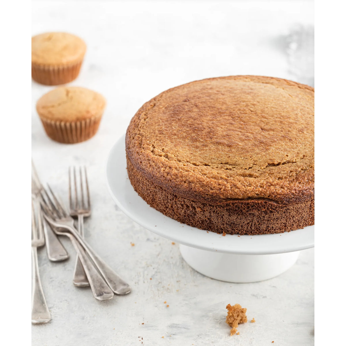 Round cake on a white stand with cupcakes and forks on a light surface