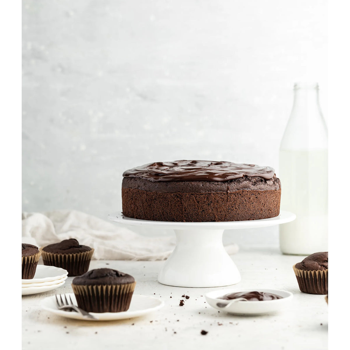 Rich chocolate cake displayed on a cake stand with a glass bottle of milk and a folded tea towel in the background, with frosted cupcakes and icing in the foreground on a clean white surface.