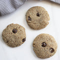 Three chocolate chip cookies on a white surface with a gray cloth in the background.