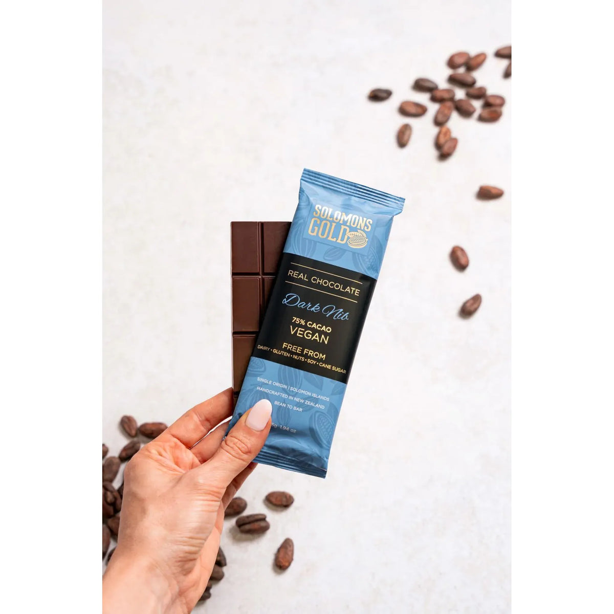 Hand holding a Solomon's Gold dark chocolate bar with a blue wrapper on a white background with scattered cacao beans.
